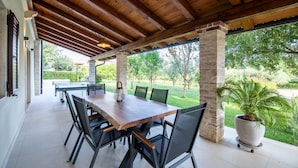 An outdoor covered patio with a wooden table and chairs, a potted plant, and a view of a green landscape. - Fiorini in Fiorini Brtonigla (Brtonigla)