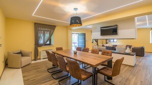 A modern living room with a large wooden dining table, comfortable seating, a flat-screen TV mounted on the wall, and a kitchen area in the background. - Fiorini in Fiorini Brtonigla (Brtonigla)