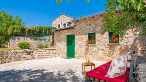 A stone building with a green door and windows with flowers. There is a red bench with a cushion and a small table with a bottle and glasses. - Ognjistar in Zmijavci (Zmijavci)