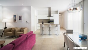A modern living room with a pink sofa, a beige sofa, and a glass dining table with a blue bowl. There is a kitchen area with a white countertop and a hanging light fixture. - Acadia C in Slatine With 6 Bedrooms and 6 Bathrooms (Split)