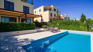 A swimming pool with lounge chairs, a covered patio area, and modern buildings in the background. - Acadia C in Slatine With 6 Bedrooms and 6 Bathrooms (Split)