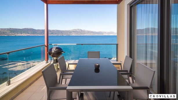 A balcony with a table and chairs, overlooking a coastal view with a grill and a clear sky. - Acadia C in Slatine With 6 Bedrooms and 6 Bathrooms (Split)