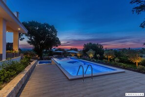 Piscina com deck de madeira, rodeada por jardins ajardinados e árvores, sob um céu crepuscular.