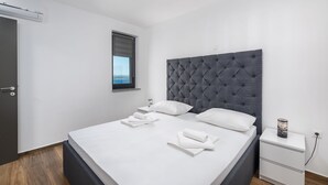 A neatly made bed with white linens and a grey tufted headboard, a bedside table with a lamp, and a window with a view. - Twins III in Crikvenica (Crikvenica)