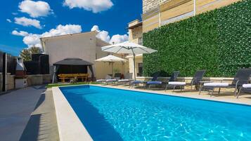 A swimming pool with lounge chairs and umbrellas, a stone wall, and a barbecue area.