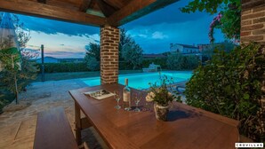 Eine Terrasse im Freien mit einem Holztisch, einer Weinflasche und Gläsern, mit Blick auf einen Swimmingpool und ein Backsteinhaus.