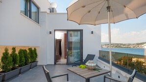 A balcony with a table, chairs, and an umbrella. There is a view of the sea and a building in the background. - More in Okrug Gornji (Okrug)