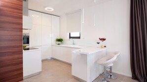 A modern kitchen with white cabinetry, a central island with bar stools, a built-in oven, and a large window. - More in Okrug Gornji (Okrug)