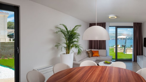 A modern dining area with a wooden table, white chairs, a potted plant, and a view of a swimming pool and cityscape through large glass doors. - More in Okrug Gornji (Okrug)