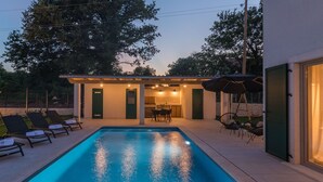 Piscina com espreguiçadeiras, área de refeições ao ar livre coberta e um edifício com porta de vidro.