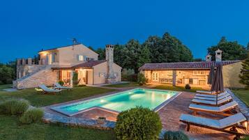Zona junto a la piscina con sillones de descanso y una pileta, rodeada por un patio de piedra y un césped cuidado.