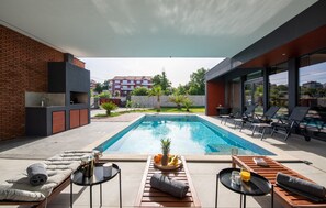 Uma área moderna com piscina ao ar livre, cadeiras de descanso, mesa de jantar com bandeja de frutas e vista para um edifício residencial ao fundo.