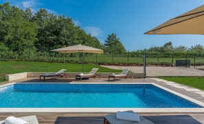 Una piscina con sillones de descanso y una sombrilla, rodeada de un jardín ajardinado y una valla.