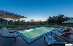 Una piscina con sillas de descanso y sombrillas, rodeada por una terraza y una zona ajardinada.