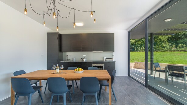 A modern kitchen with dark cabinets, a wooden dining table, and blue chairs. - Complex Teca in Go an Draguzeti (Barban)
