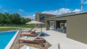 A modern outdoor pool area with lounge chairs and a table, surrounded by a house with large windows and a covered patio. - Complex Teca in Go an Draguzeti (Barban)