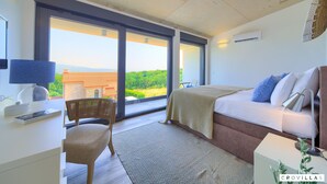 Ein modernes Schlafzimmer mit einem großen Bett, einem Schreibtisch mit Lampe, einem Sessel und Blick ins Freie durch große Fenster.