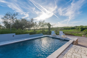 A swimming pool with a stone border, two white lounge chairs, and a grassy landscape with trees in the background. - Pendolina in Buje With 3 Bedrooms and 1 Bathrooms (Buje)