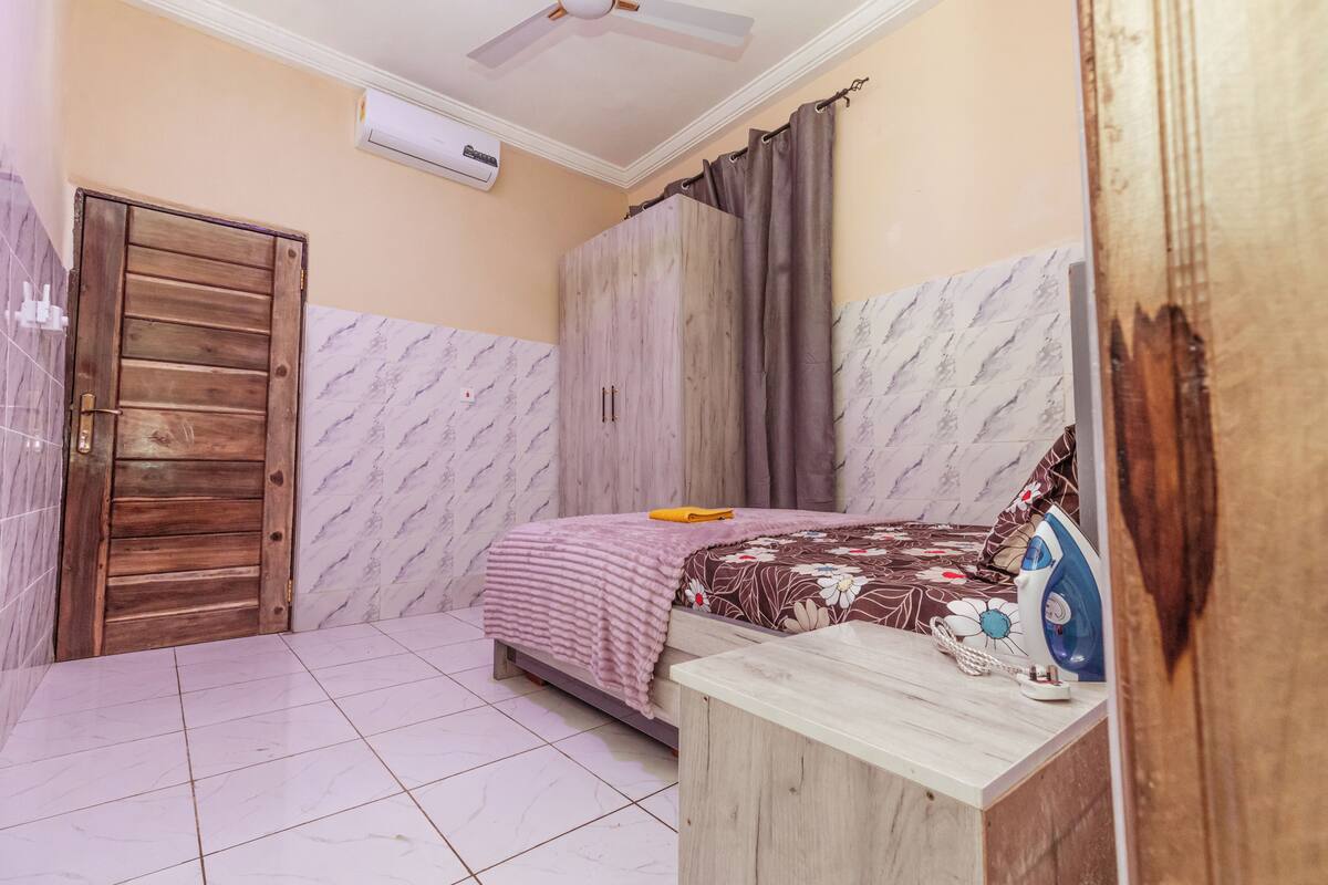 A bedroom with a bed, a wooden wardrobe, a bedside table with an iron, and a ceiling fan.