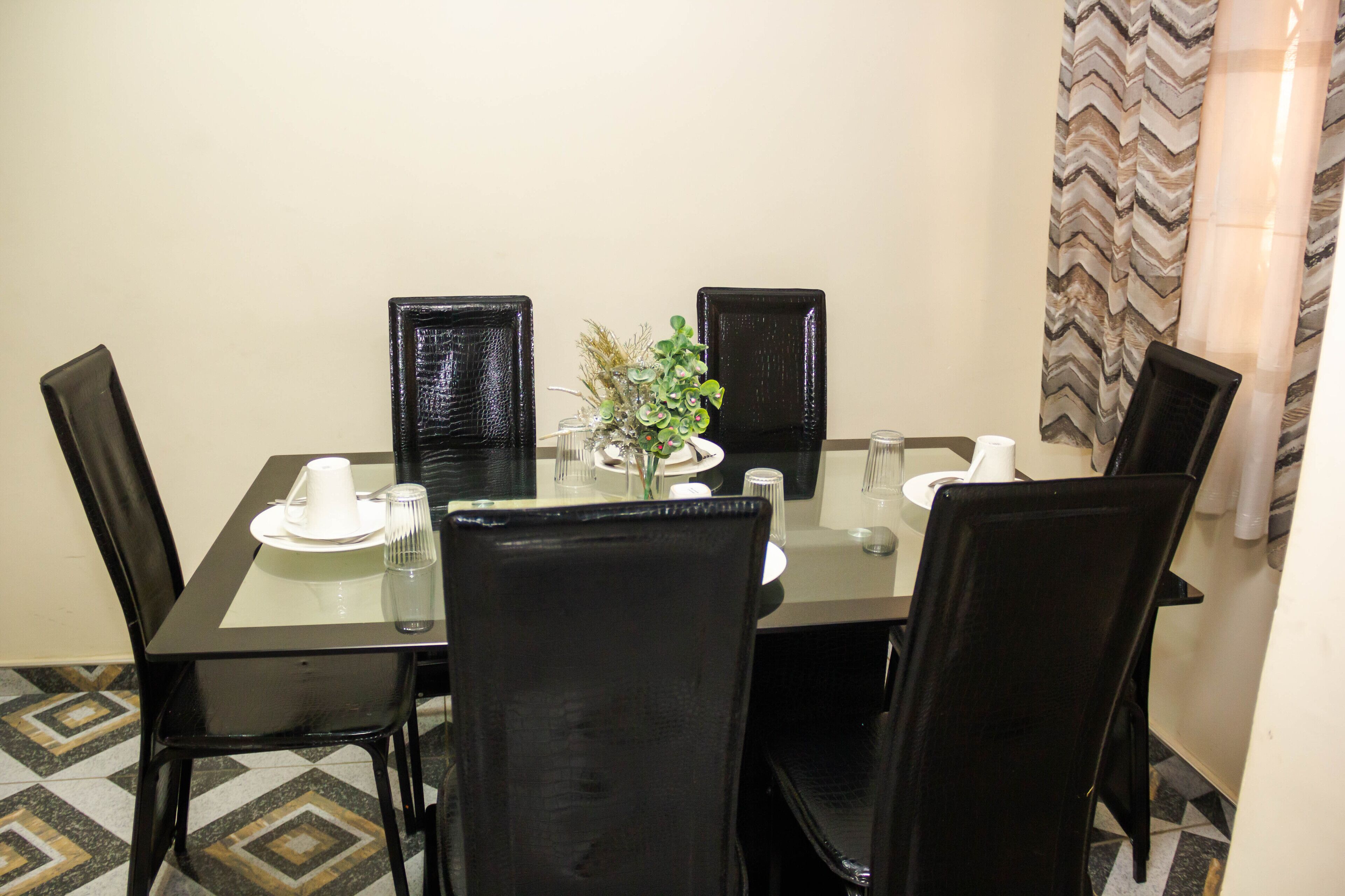 Uma área de refeições com mesa de vidro, cadeiras pretas e um vaso com flores.