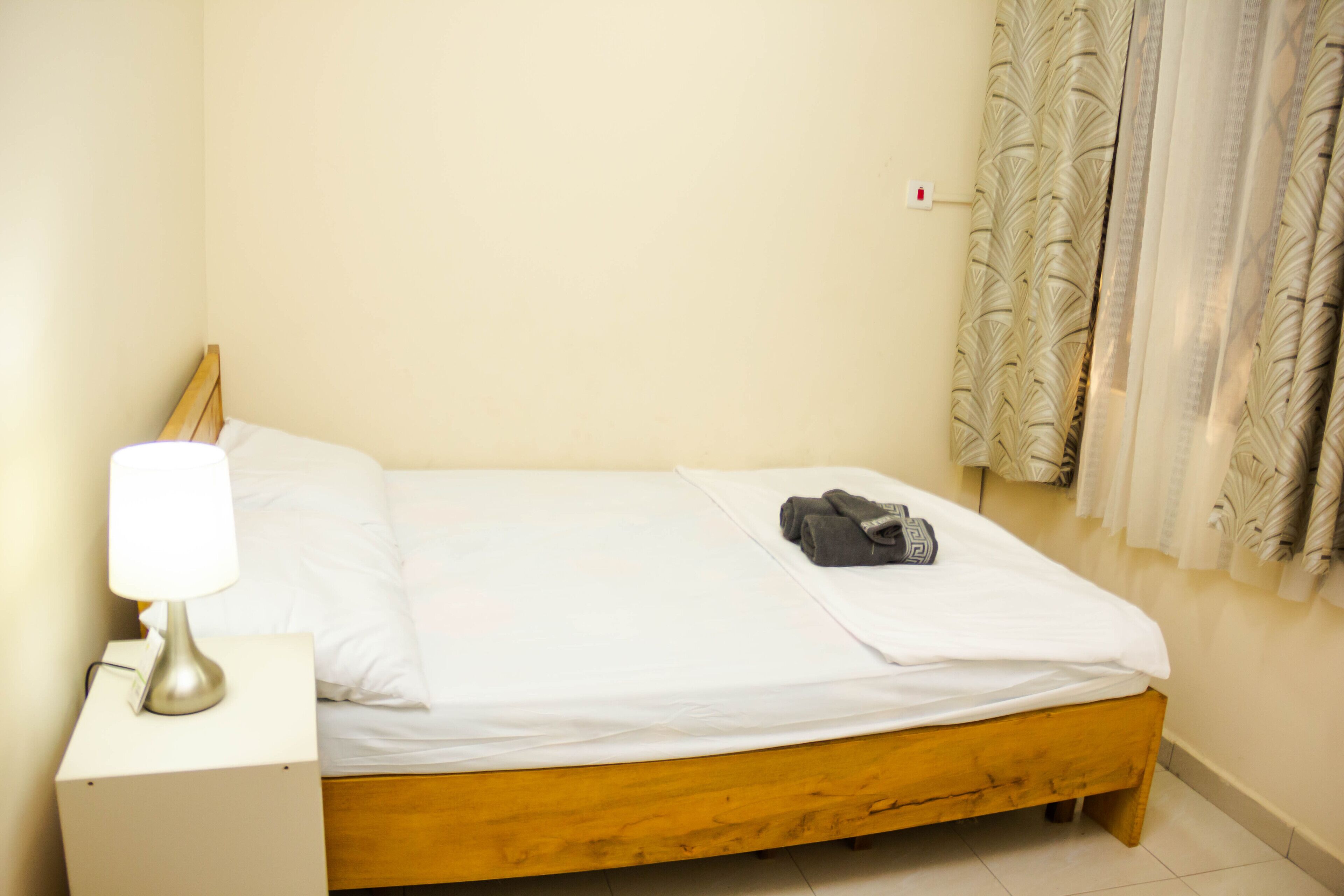 A small, neatly made bed with a wooden frame and white bedding. There is a lamp on a bedside table to the left.