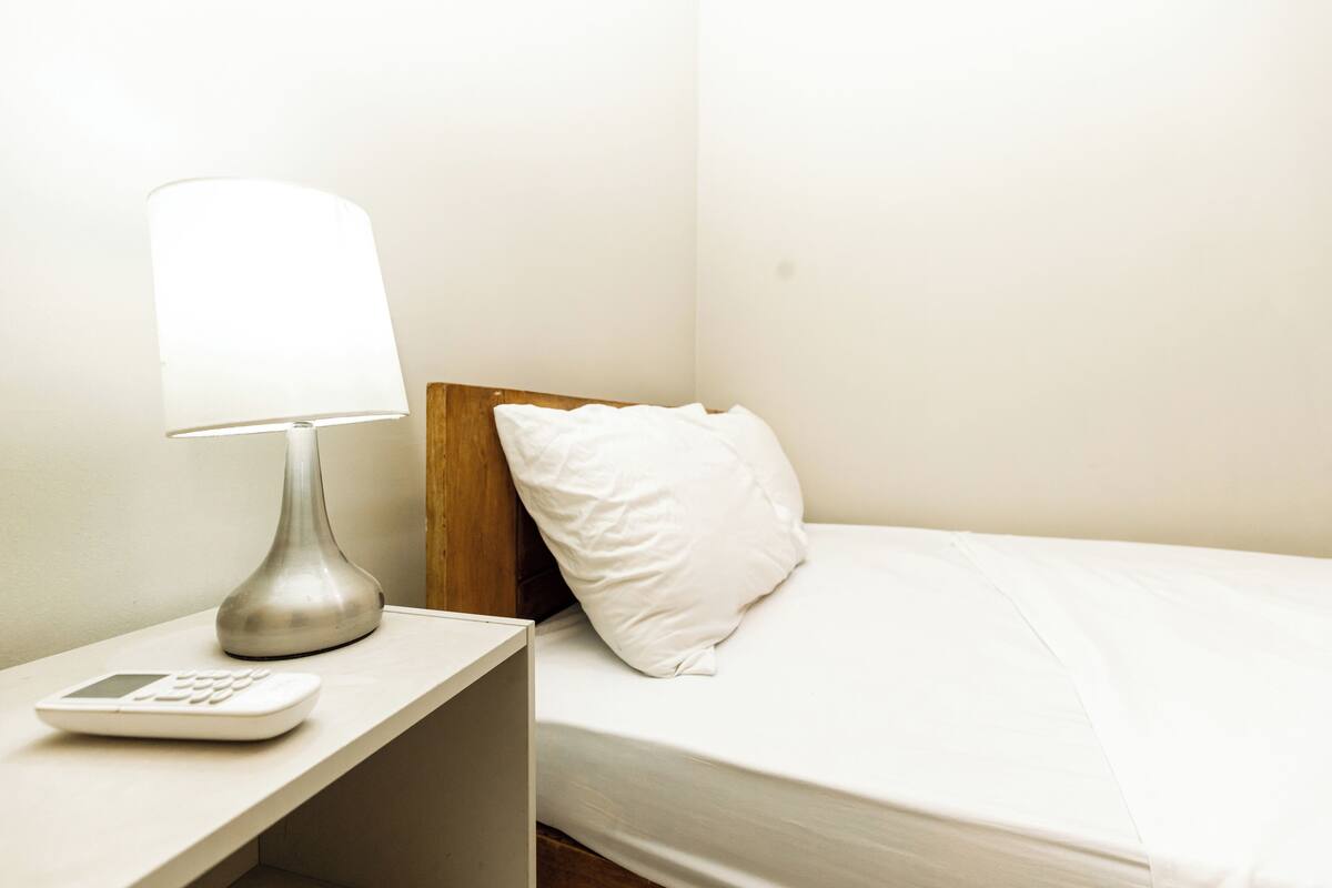 A bed with a white pillow, a bedside table with a lamp, and a small electronic device.