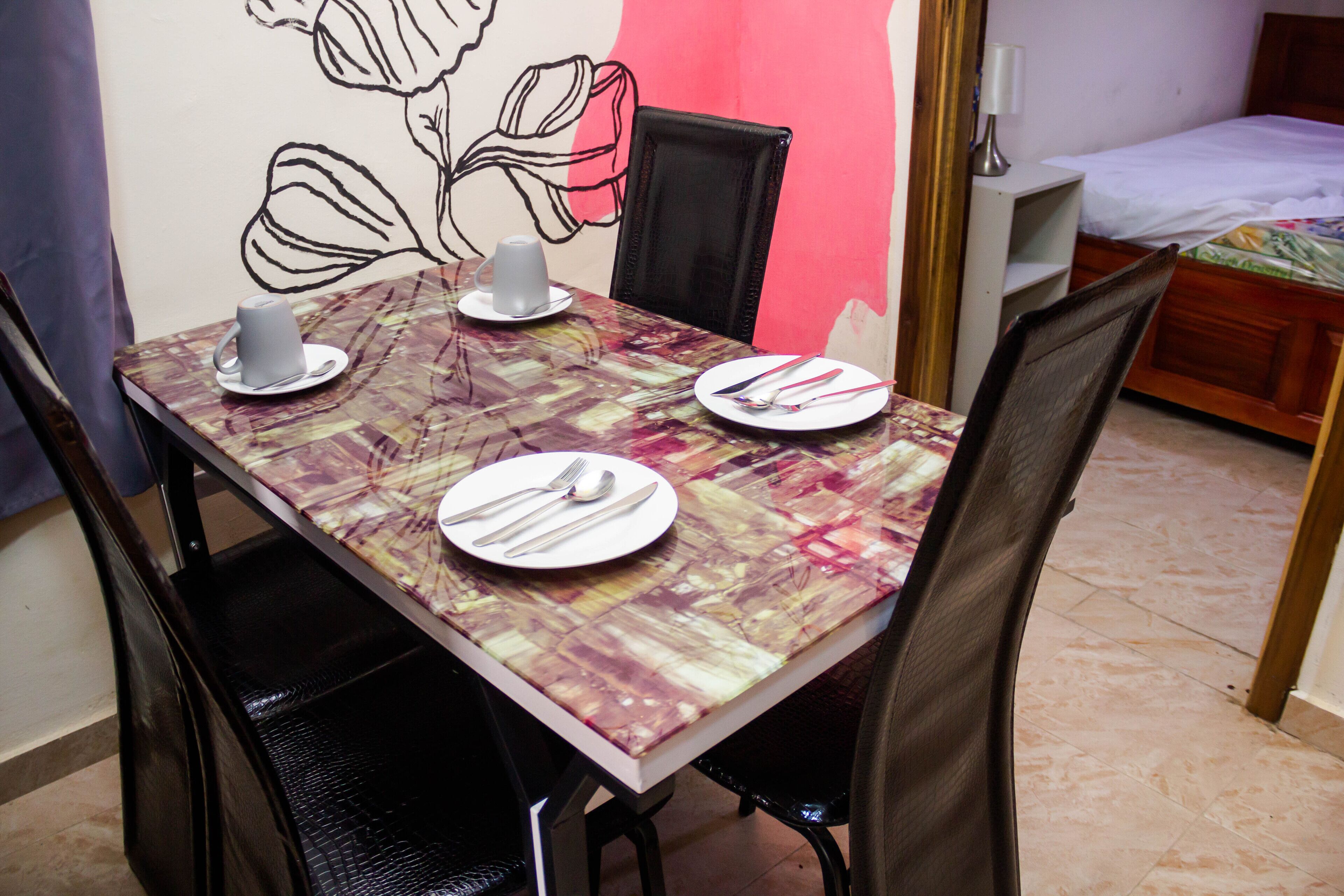 A dining table set with plates, cutlery, and cups, surrounded by chairs in a room with a bed and a wall with a floral design.