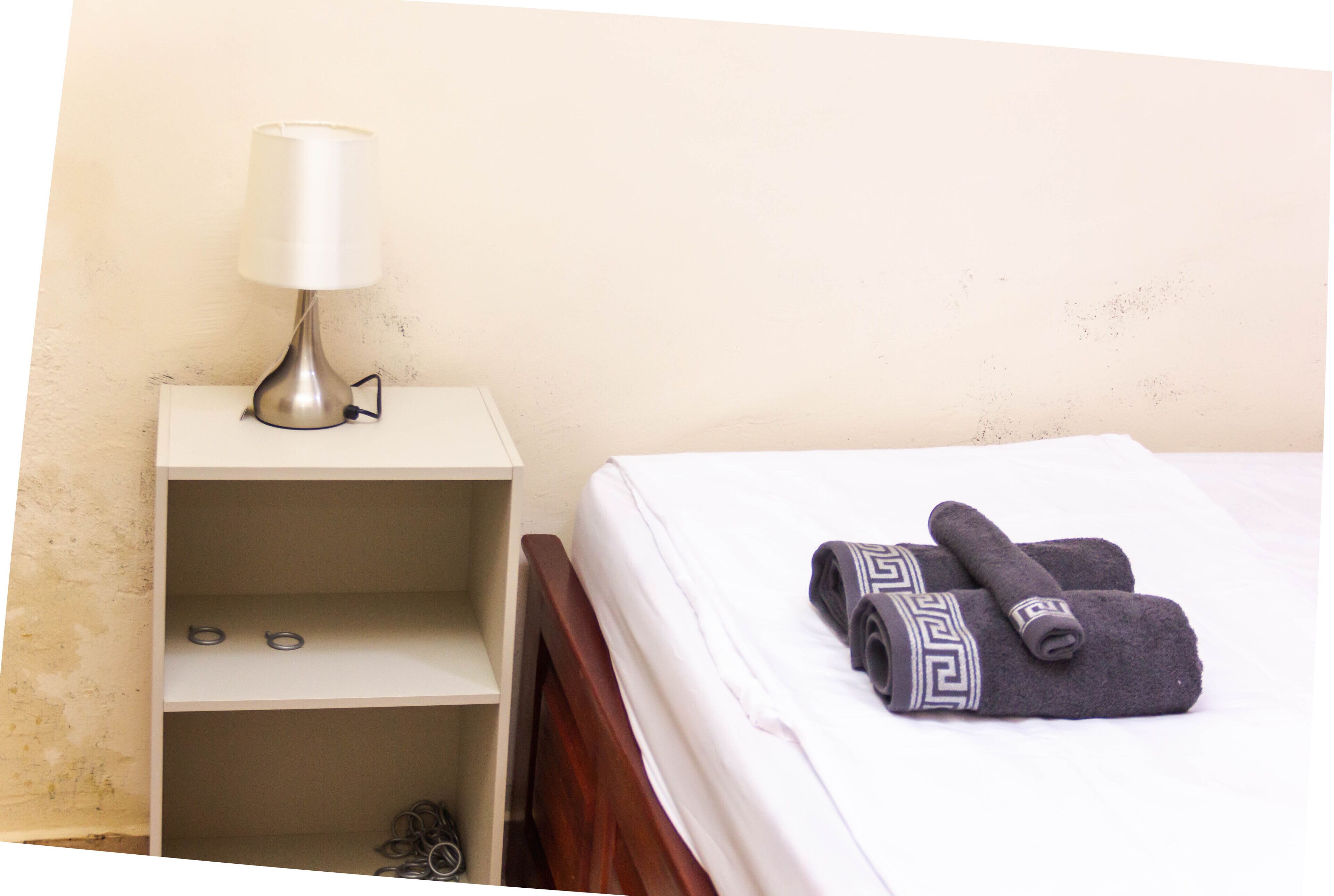 A neatly made bed with rolled towels, a bedside lamp, and a shelf with a few items.