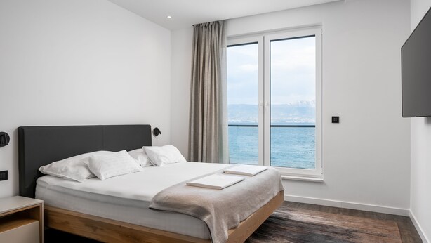Een moderne slaapkamer met een houten bed, nachtkastje, groot raam met uitzicht op zee en een wandgemonteerde televisie.