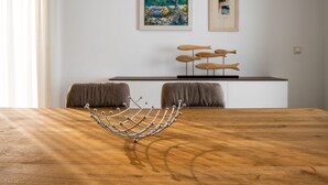 Een houten tafel met een metalen decoratief element in het midden, een witte muur met twee ingelijste kunstwerken, en een witte console tafel met enkele spullen erop.