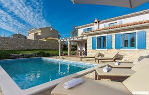 En swimmingpool med solstolar och en pergola, omgiven av stenmurar och en byggnad med blåa fönsterluckor.