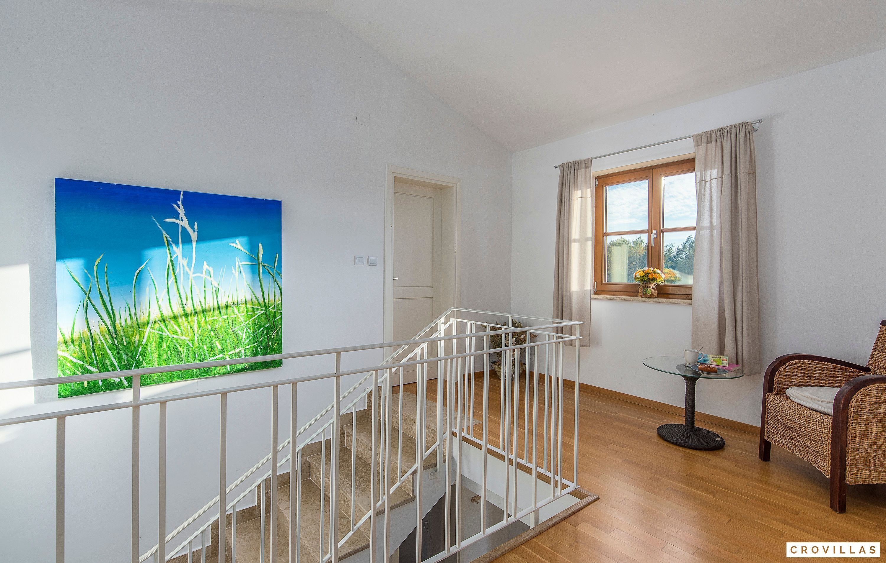 A staircase with a white railing, a painting of grass on the wall, a window with curtains, and a wicker chair.