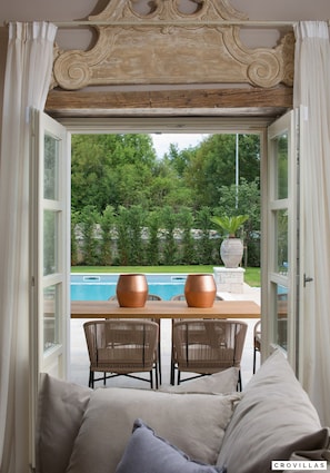Abre-se uma porta para uma área de piscina com cadeiras de vime e uma mesa, tudo envolto em vegetação.