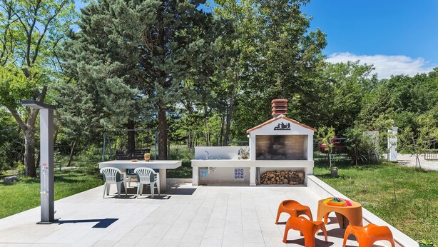Un espace cuisine extérieur comprenant un barbecue, une table avec des chaises et un petit coin salon avec des tabourets orange.
