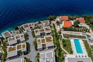 An aerial view of a coastal hotel with multiple buildings, a swimming pool, and a clear view of the sea.