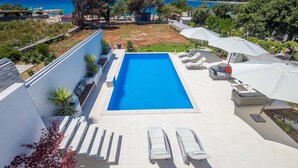 A rectangular swimming pool with lounge chairs and umbrellas, surrounded by greenery and a view of the sea. - Andreja in Punat With 6 Bedrooms and 6 Bathrooms (Punat)