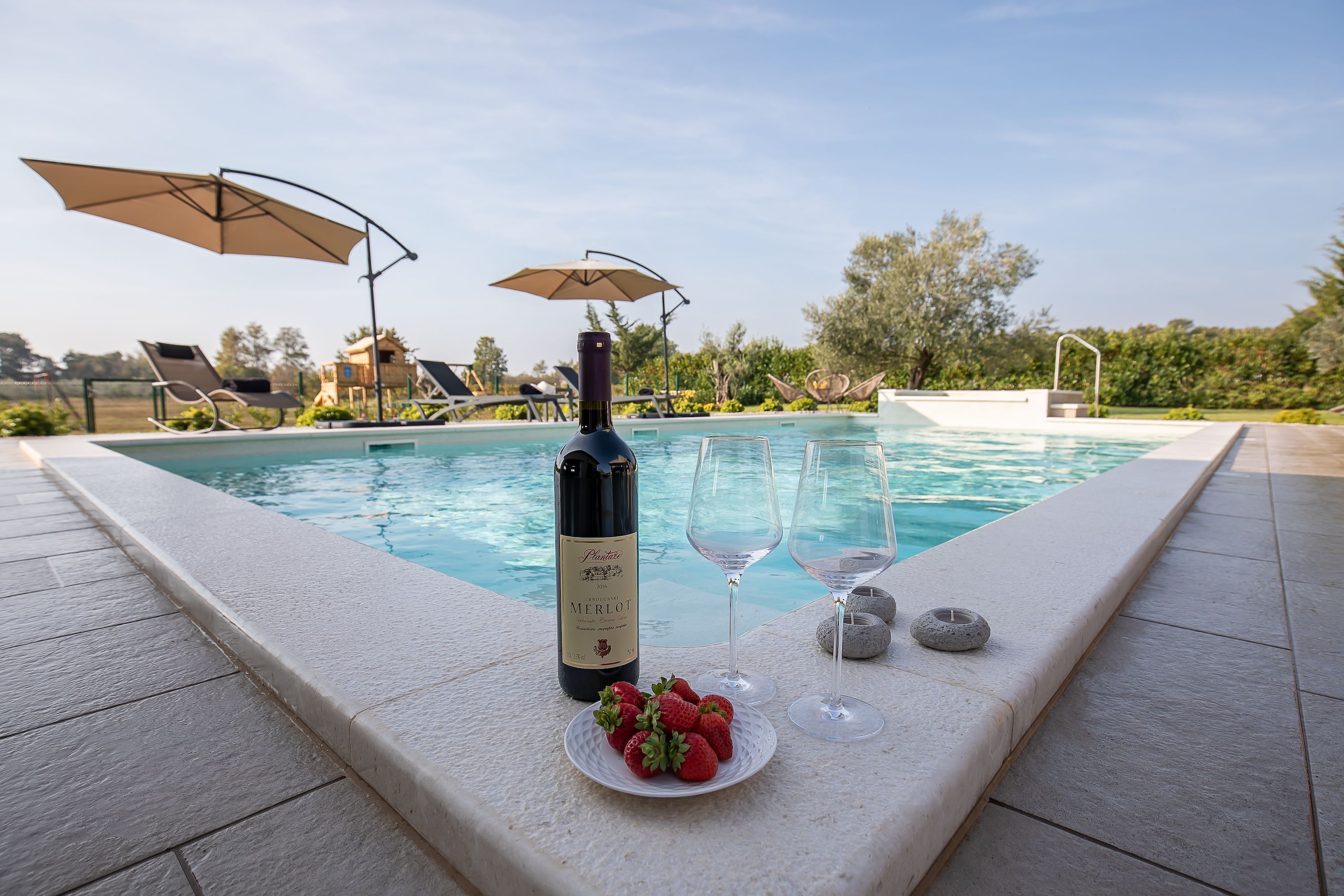 Escena junto a la piscina con una botella de vino Merlot, dos copas y un plato de fresas.