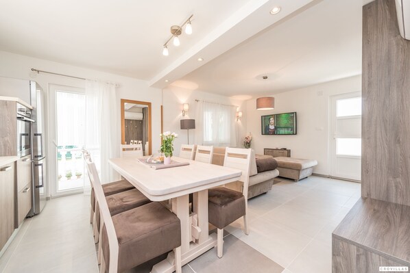 Eine moderne Küche mit Einbaugeräten, ein Essbereich mit weißem Tisch und braunen Stühlen sowie ein Wohnbereich mit Sofa und Fernseher.