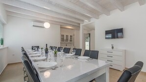 A dining area with a long table set for six, white walls, a ceiling with exposed beams, a television mounted on the wall, and a kitchen area in the background. - Oliva in Premantura With 6 Bedrooms and 6 Bathrooms (Medulin)
