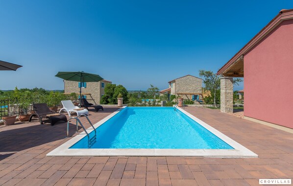 Uma piscina retangular com superfície de água azul cristalina, rodeada por um deck pavimentado. Há espreguiçadeiras e um guarda-sol coberto no deck.