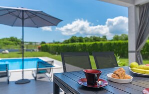 En terrasse med swimmingpool, udendørs møbler og et bord med croissant og frugt.