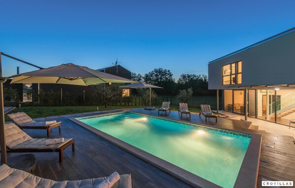 A modern outdoor pool area with lounge chairs and umbrellas, surrounded by a wooden deck and a glass-enclosed building. - Caeli in Go an Draguzeti (Barban)