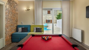A pool table in a room with a stone wall, a green sofa, and a view of an outdoor pool area. - Loki in Vi Inada With 4 Bedrooms and 4 Bathrooms (Vizinada)