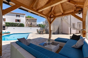 A covered outdoor seating area with blue cushions, a pool, and stone buildings in the background. - Loki in Vi Inada With 4 Bedrooms and 4 Bathrooms (Vizinada)
