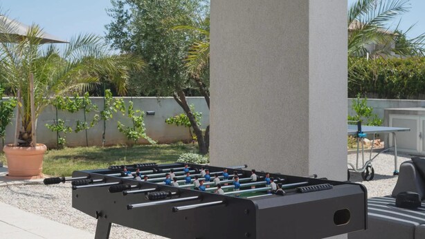 Ein Tischkicker im Freien auf einer Terrasse, neben einer Betonstütze, umgeben von Grünpflanzen und einer weißen Wand.