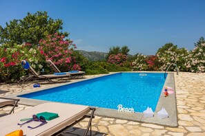 Une piscine entourée de chaises longues, d’une terrasse en pierre et d’arbustes à fleurs roses.