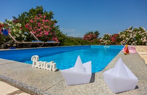 Une piscine avec un panneau indiquant « Plage », des chaises longues et des fleurs roses en arrière-plan.