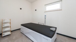 Ein Behandlungsraum mit einem Bett, einem Fenster und einer Ablage mit Handtüchern.