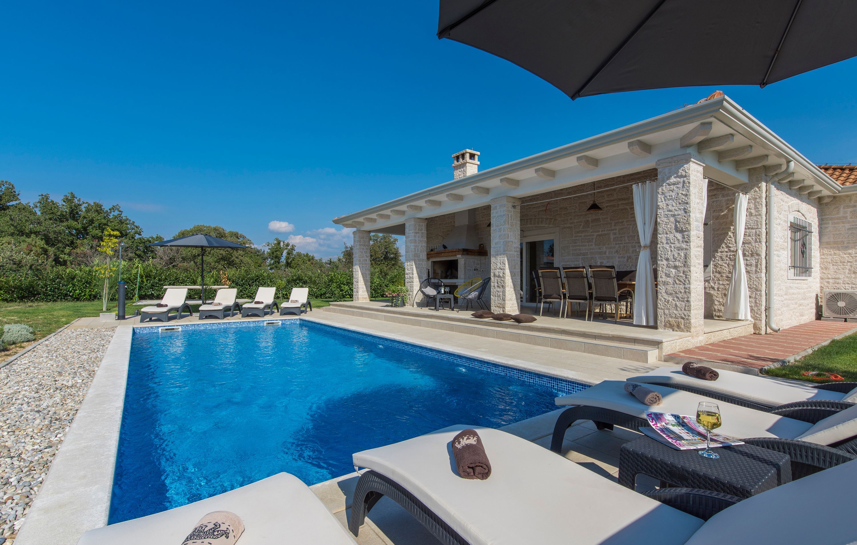 A swimming pool with lounge chairs and umbrellas, a covered patio area, and a stone building with a chimney.