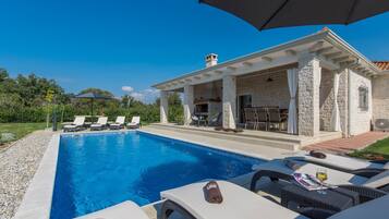 A swimming pool with lounge chairs and umbrellas, a covered patio area, and a stone building with a chimney.